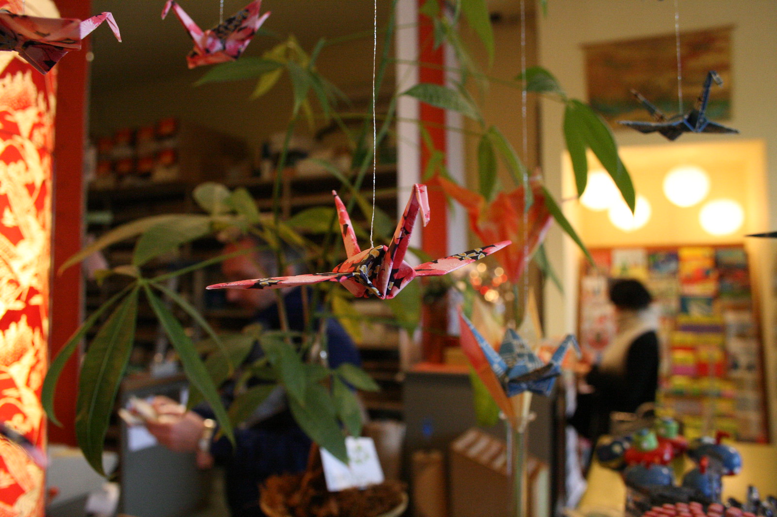 Patterned origami paper cranes hanging on strings inside a warmly lit shop with customers browsing colorful goods.