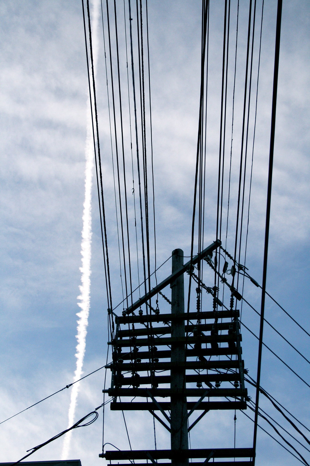 A utility pole tangled with power lines silhouetted against a cloudy sky, a vertical jet contrail streaking behind.