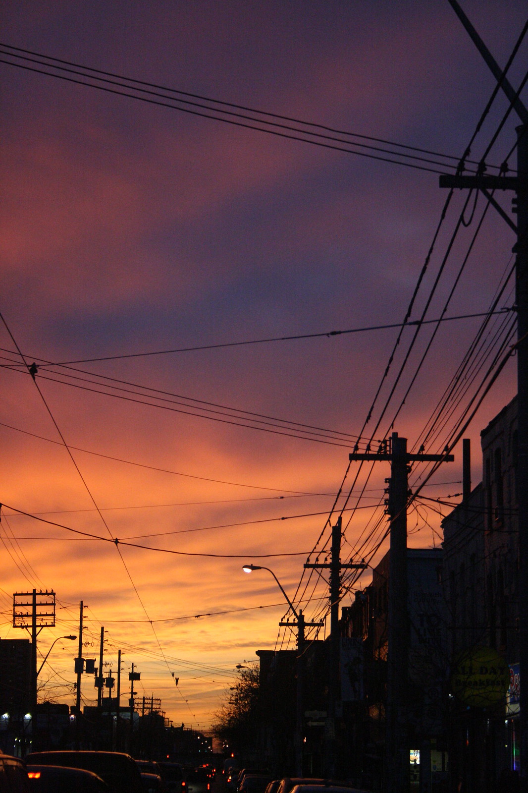 Fiery orange-and-purple sunset over a city street silhouetted by tangled utility wires, poles, and an "All Day Breakfast" sign.