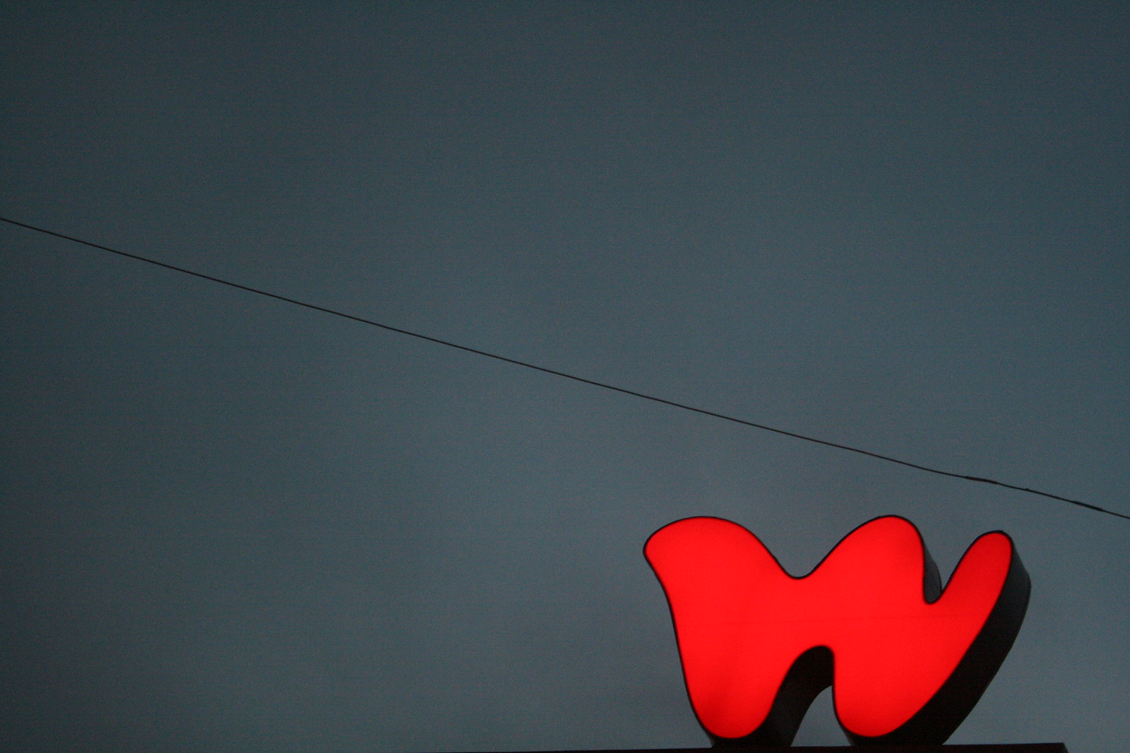 Illuminated red Woolworths "W" sign glows against a slate-grey dusk sky crossed by a power line.