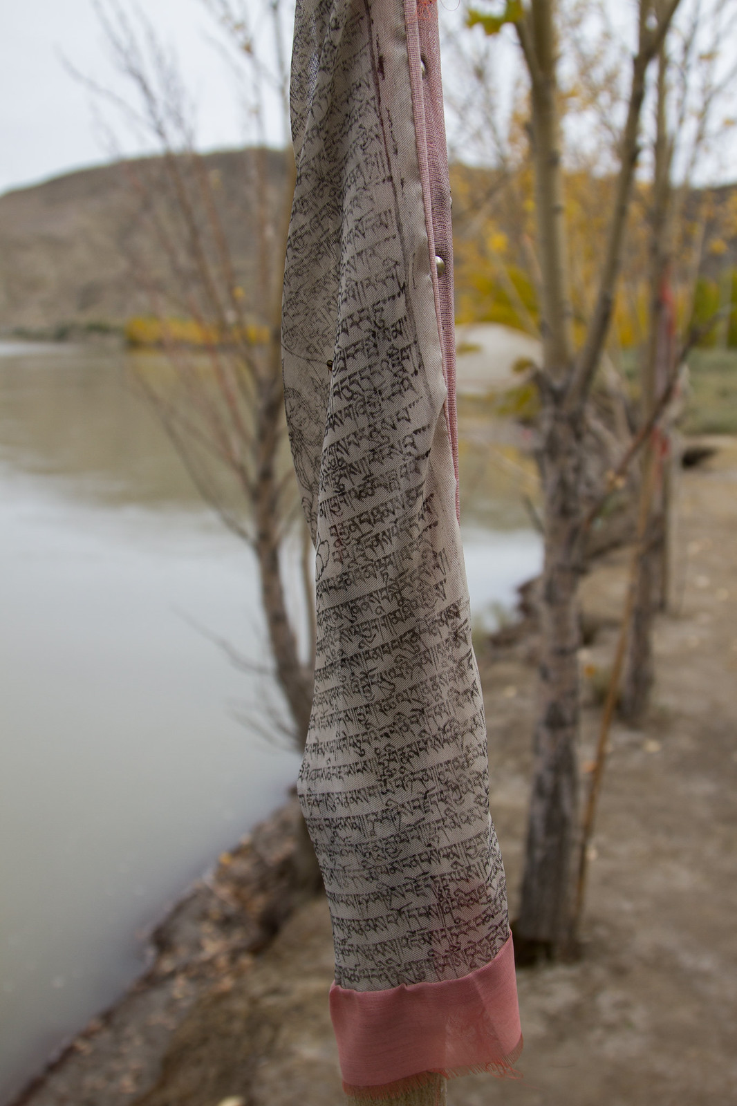 A weathered Tibetan prayer flag printed with Buddhist scriptures hangs by a calm river lined with bare autumn trees.