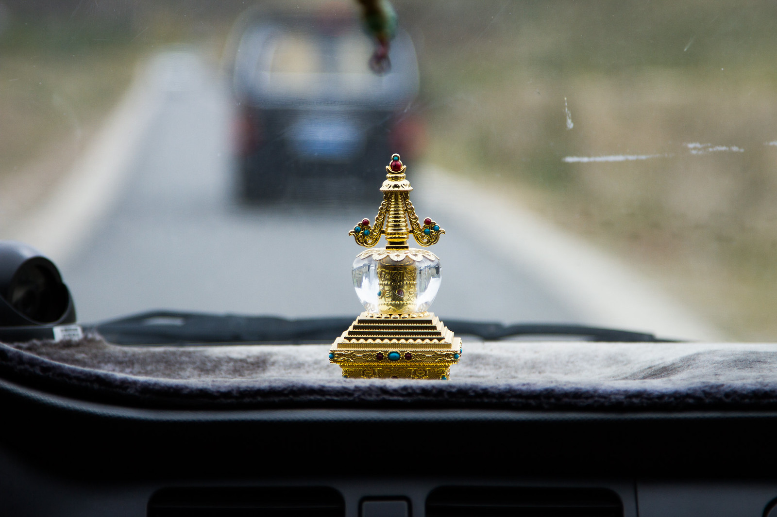 Jeweled gold and crystal Tibetan stupa ornament on a car dashboard, a vehicle ahead on the road beyond the windshield