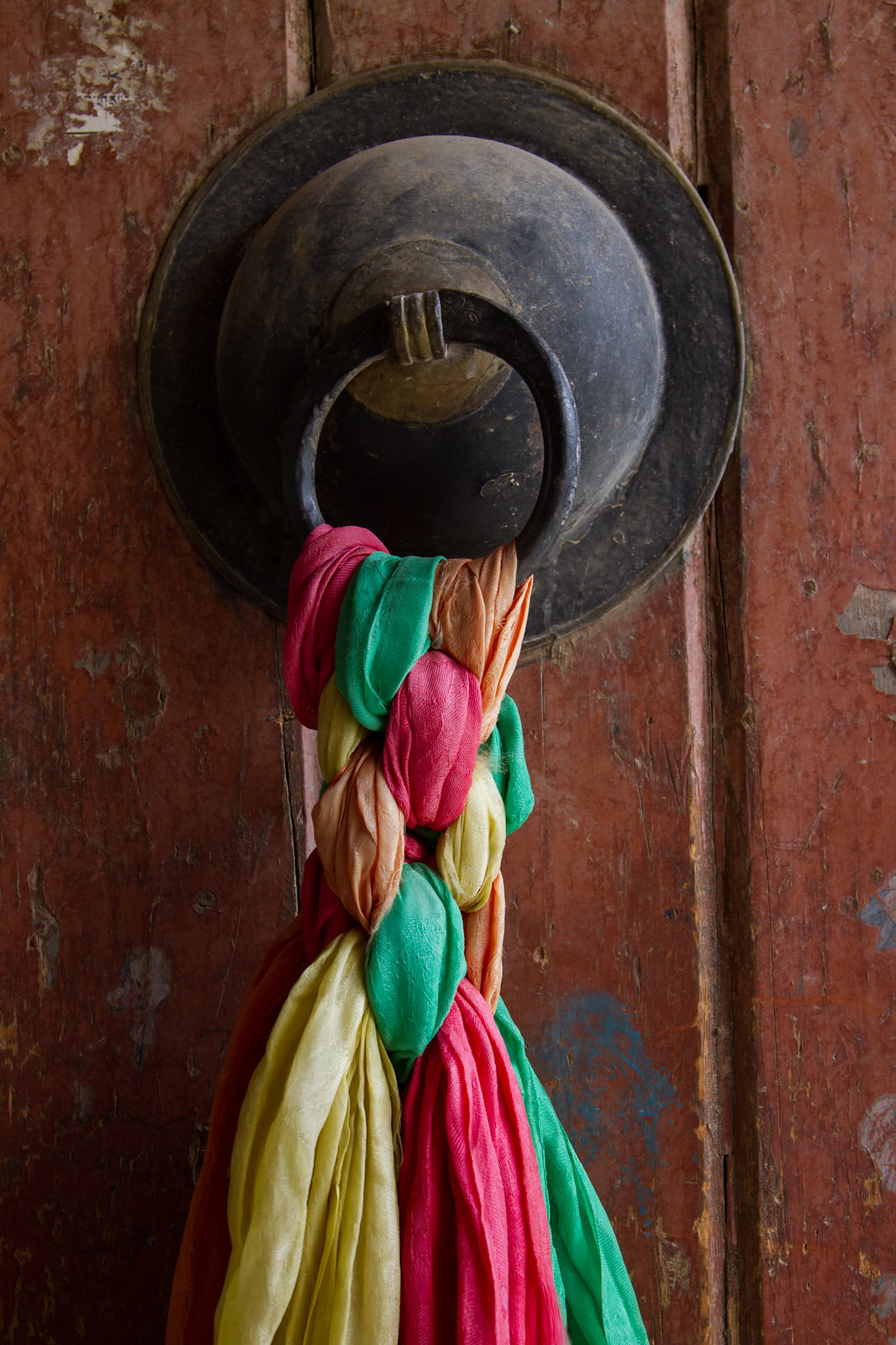 Colorful silk scarves braided through a black iron ring handle on a weathered red Tibetan door.