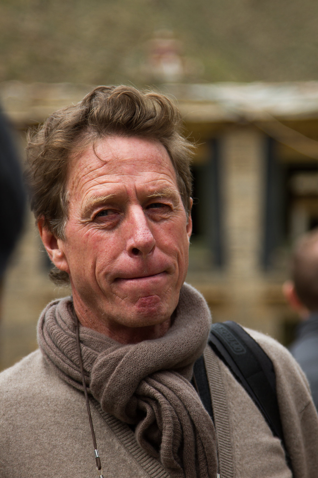 Weathered middle-aged man in a beige sweater and scarf with a backpack, standing outdoors before a building.