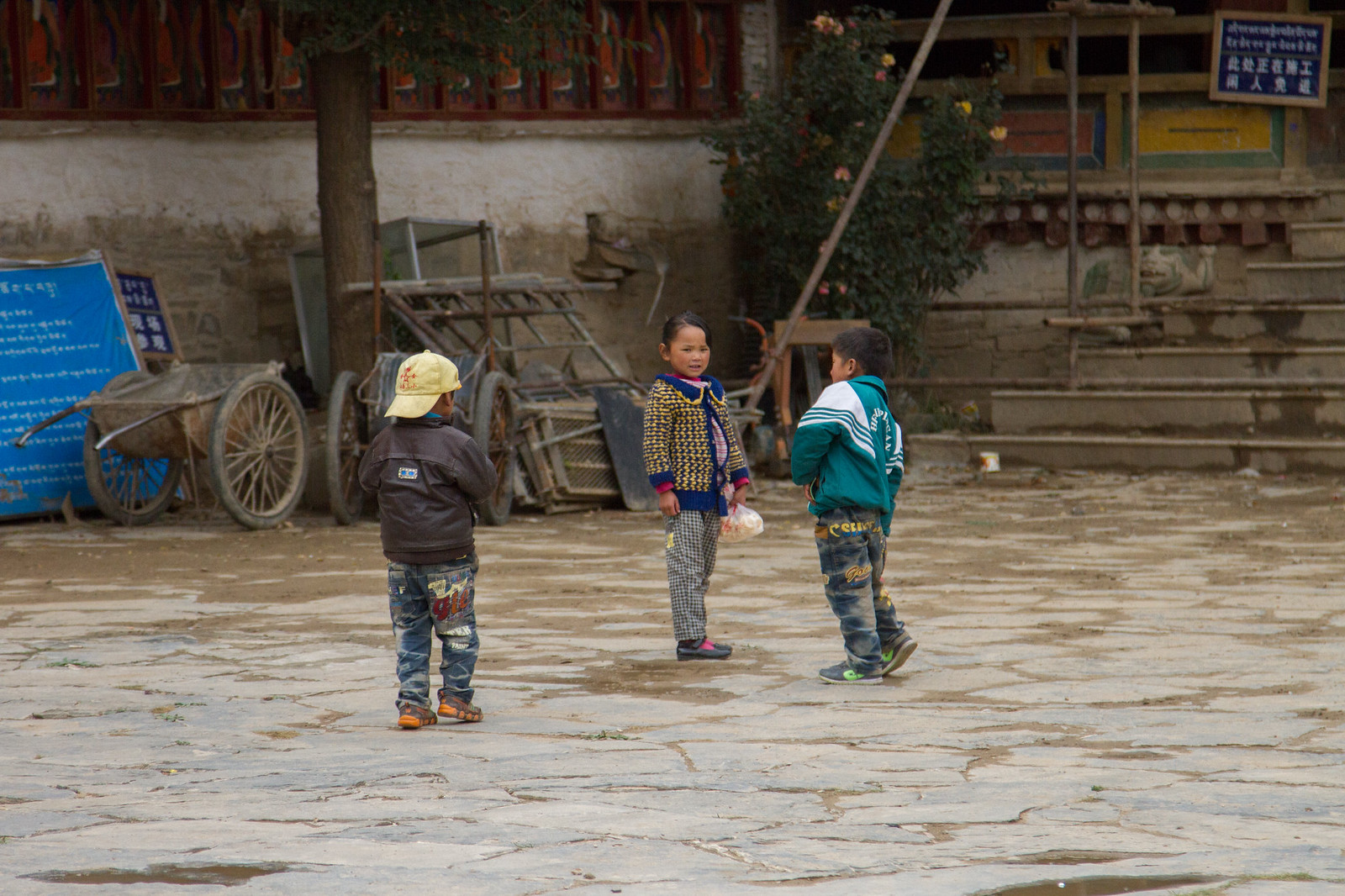 Three children play in a stone courtyard outside a Tibetan monastery with a wooden cart and painted walls.