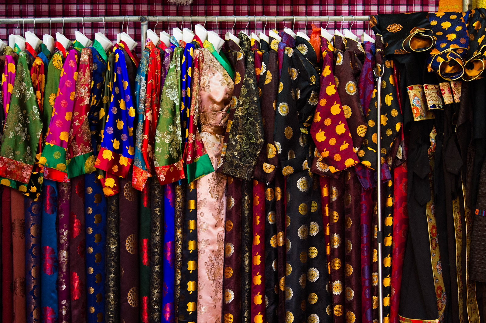 Rack of colorful Tibetan silk brocade robes and chubas hanging on white hangers at a market stall.