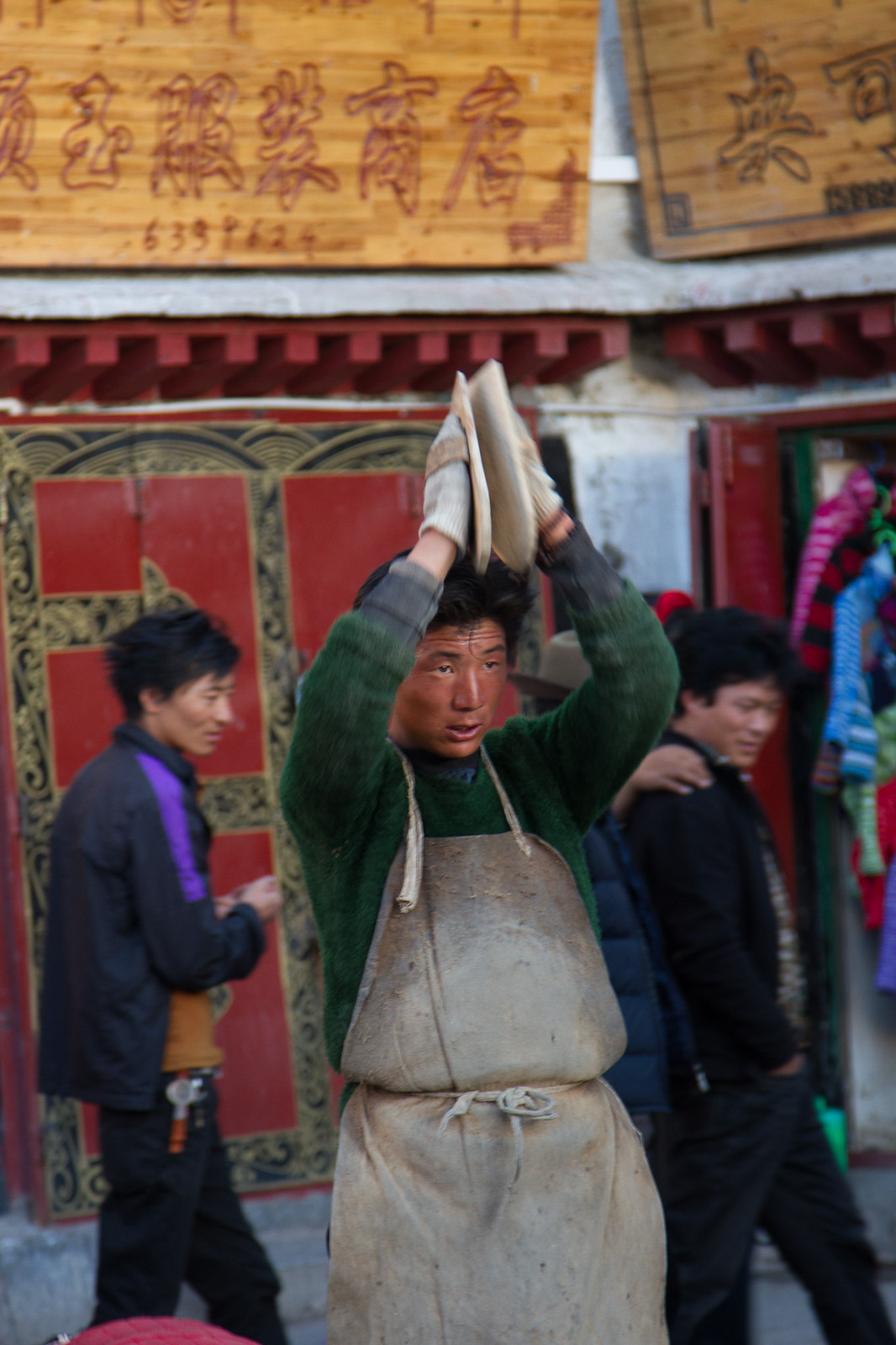 A young man in a green sweater and canvas apron carries a load atop his head past a Tibetan shop with carved wooden signs.