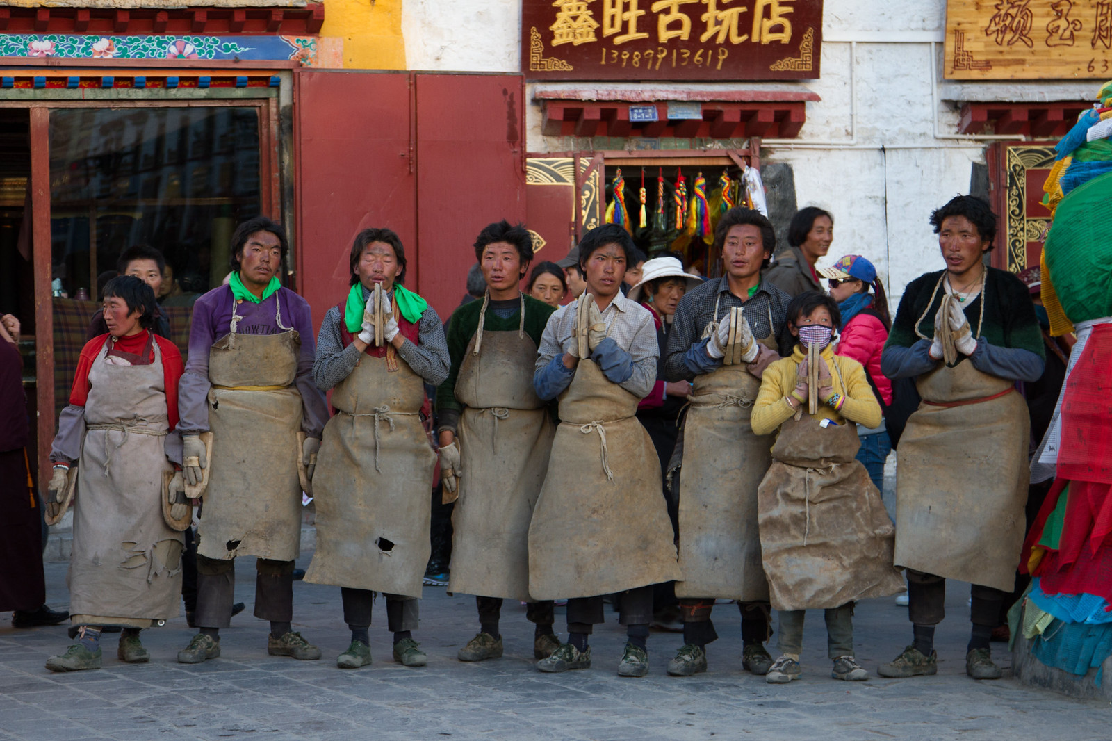 Tibetan pilgrims in leather aprons and hand guards prostrating in prayer outside shops near Lhasa's Jokhang Temple.