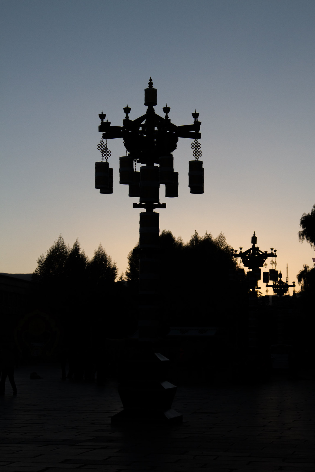 Ornate Tibetan lantern street lamps silhouetted against a dusk sky over a plaza with trees.