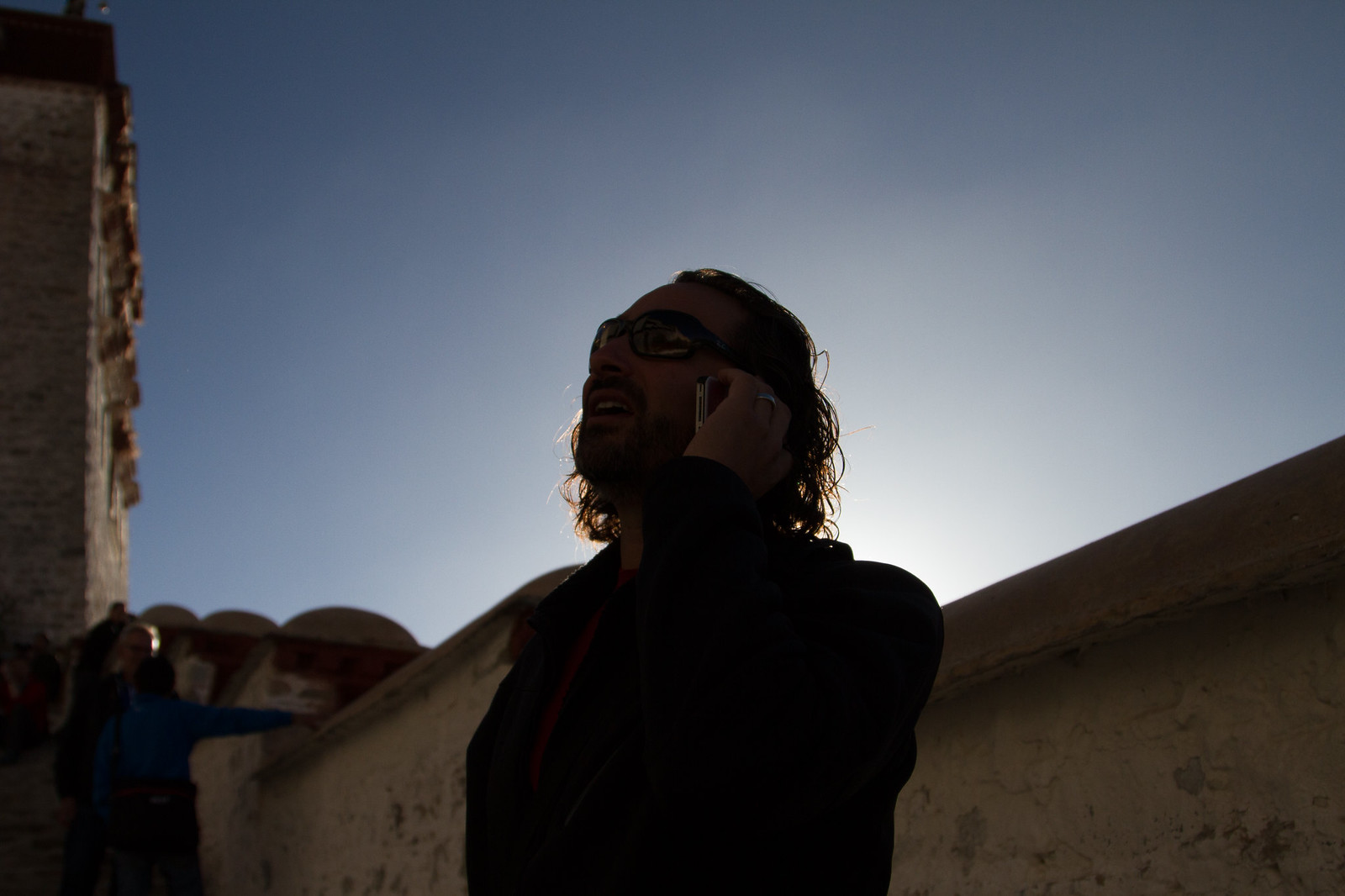 Backlit man in sunglasses talks on a phone beside a whitewashed monastery wall at dusk
