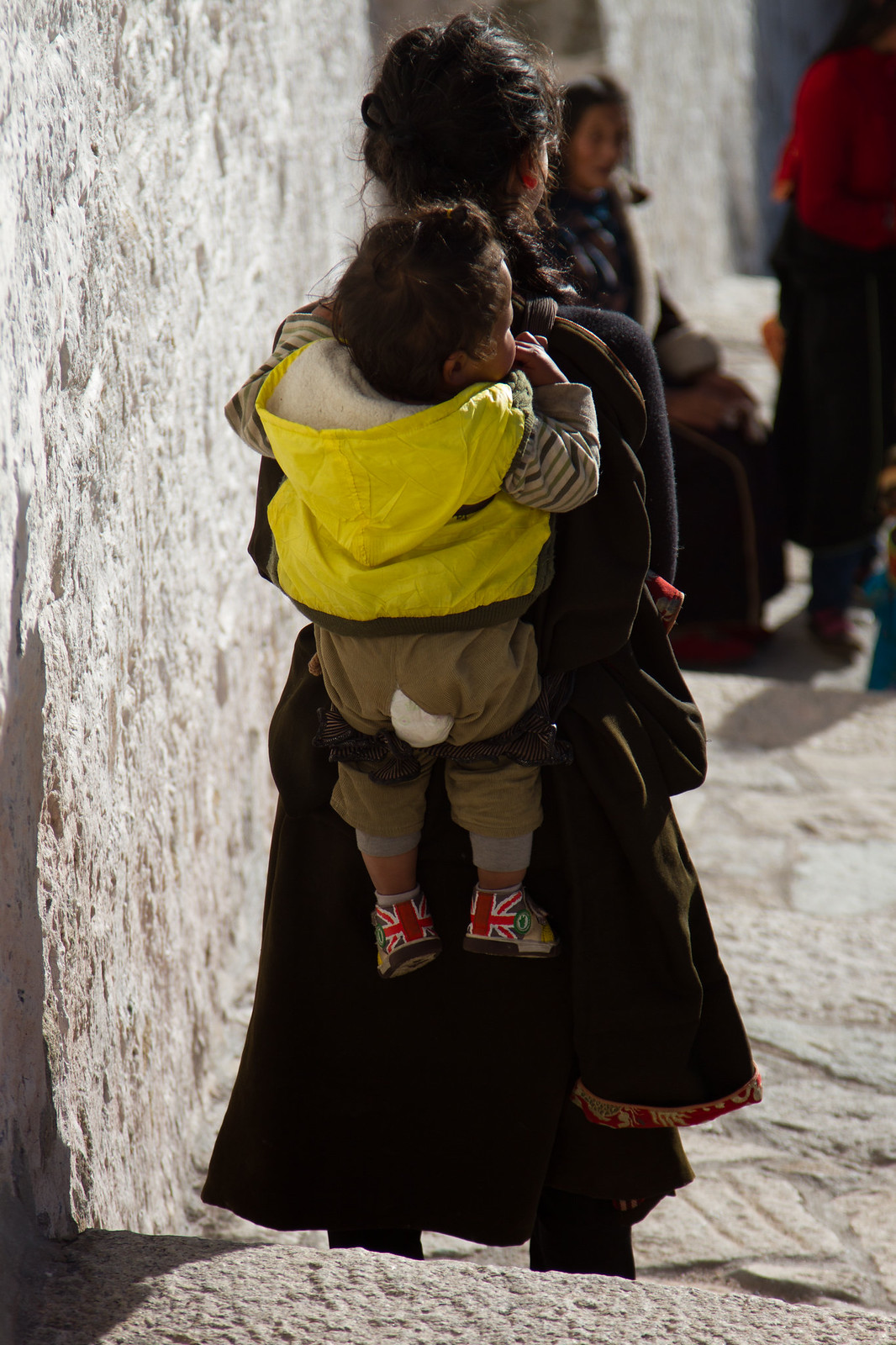 A Tibetan woman in a dark chuba carries a child in a bright yellow hooded jacket and Union Jack sneakers along a sunlit whitewashed alley.