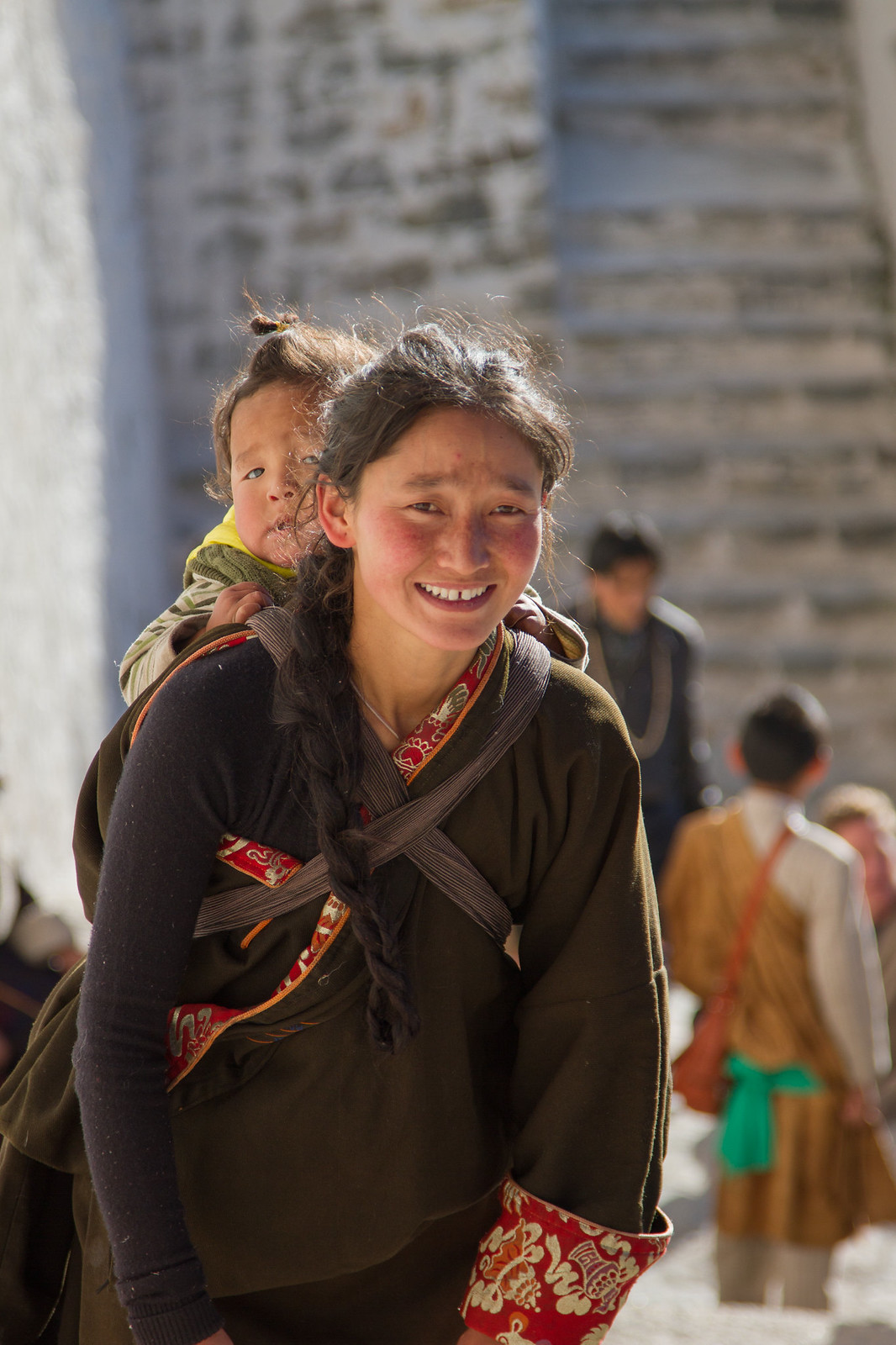 Smiling Tibetan woman with a long braid carries a child on her back in a sunlit stone courtyard.