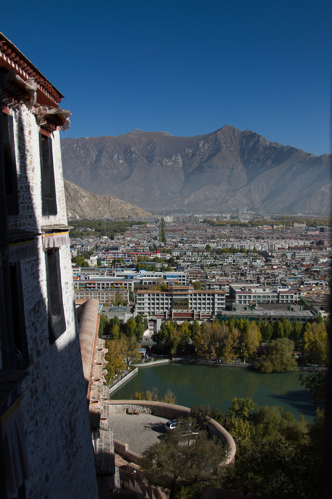 View from a whitewashed Tibetan palace wall over Lhasa's rooftops, a pond, and mountains under blue sky.