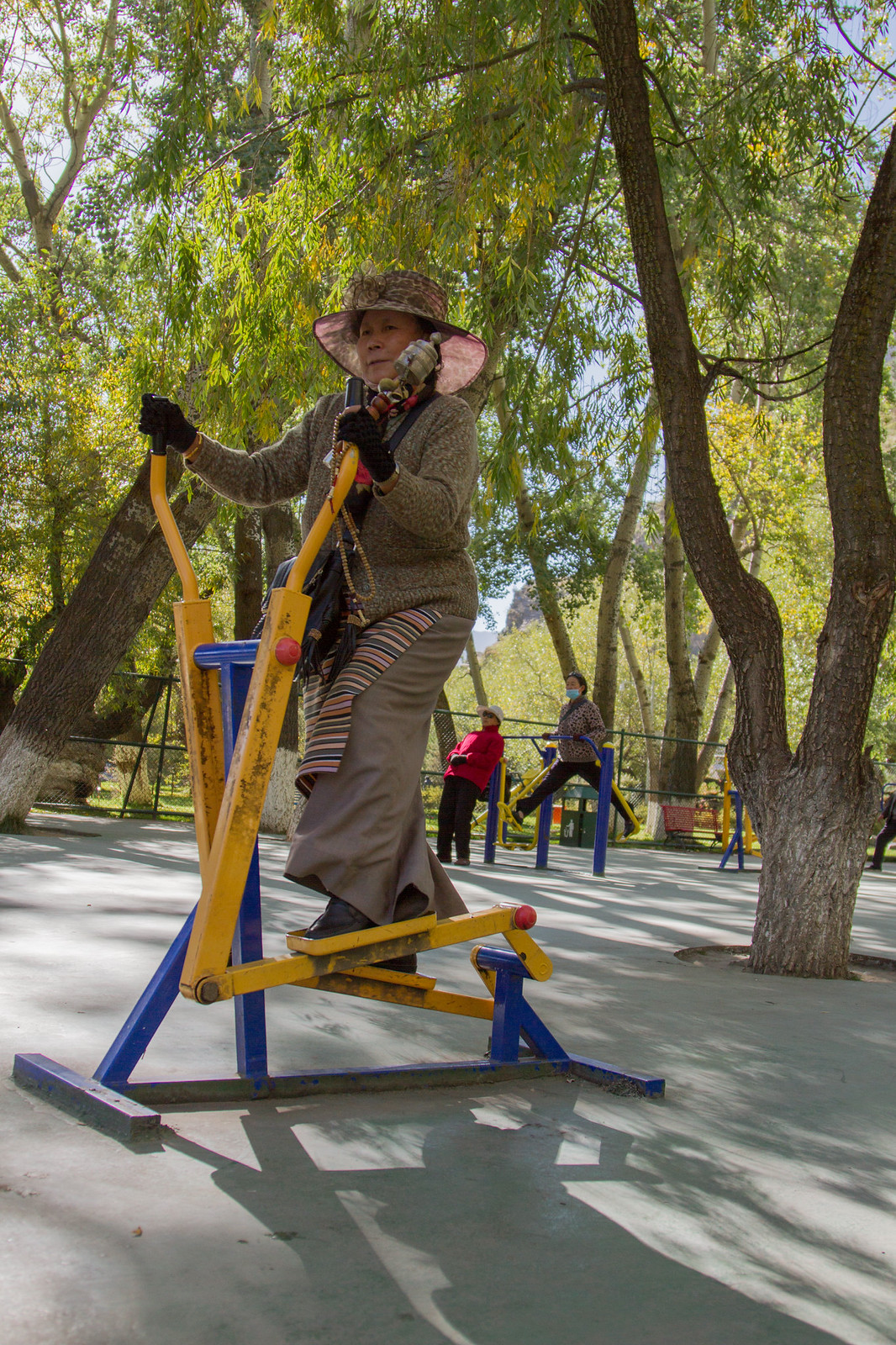 An older Tibetan woman in a sun hat and striped apron uses yellow outdoor exercise equipment in a tree-lined park.
