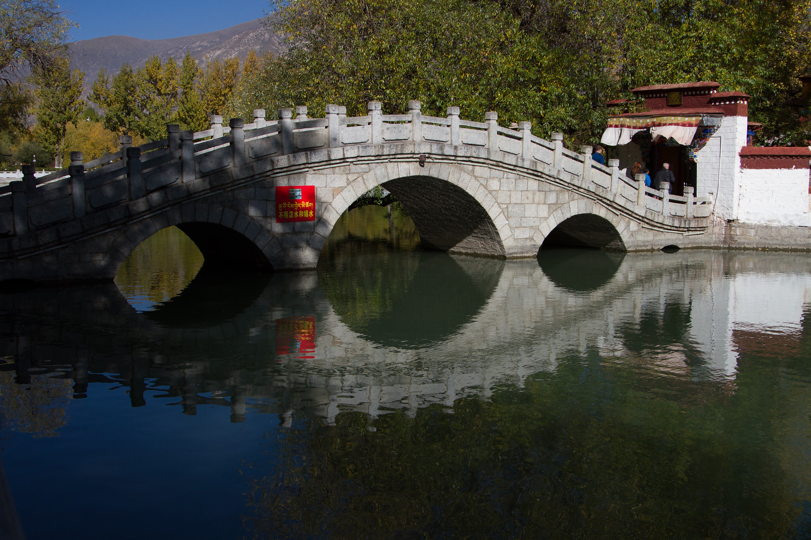 Arched white stone bridge with three arches reflecting in calm water, mountains and trees behind under a blue sky.