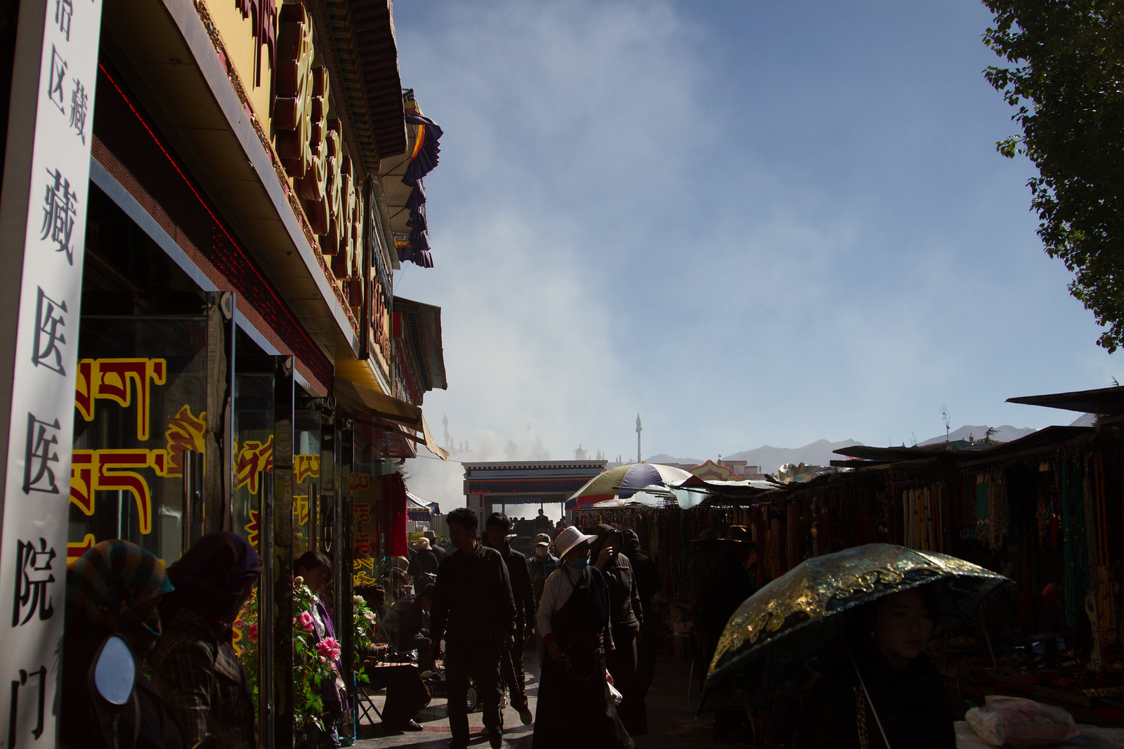Backlit Tibetan market street with vendors, umbrellas, incense smoke, and a Tibetan medicine hospital sign in Lhasa.