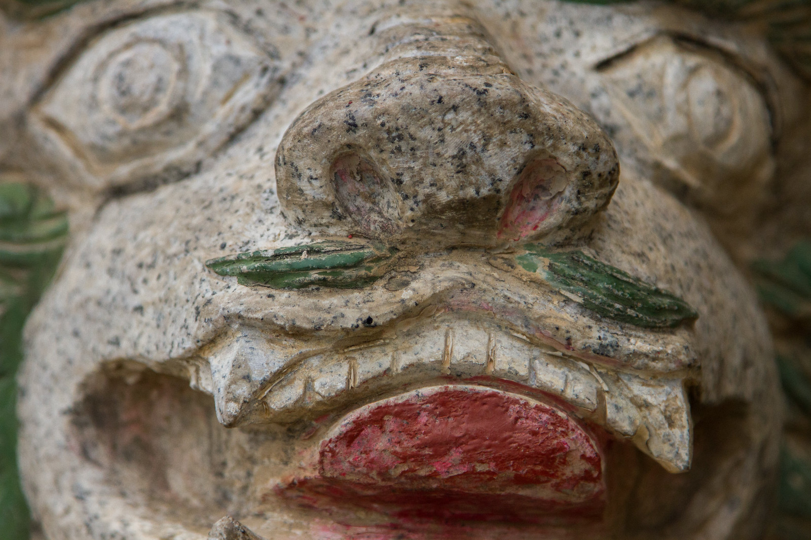 Weathered stone lion sculpture in close-up, with red-painted mouth and green-tinted whiskers.