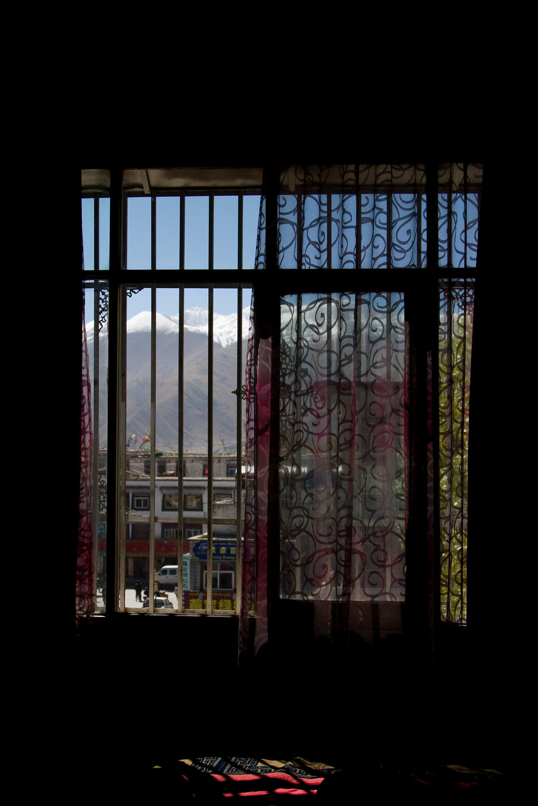 View through a barred, lace-curtained window of a Tibetan town street and snow-capped mountains beyond.
