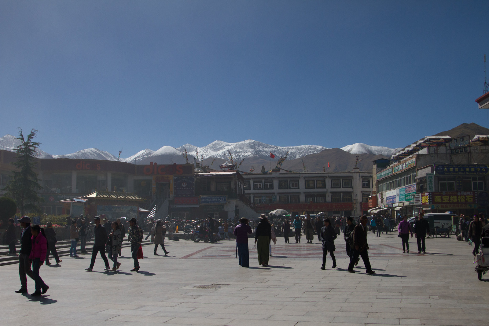 People crossing a sunlit Lhasa plaza lined with shops, backed by snow-capped Himalayan peaks under blue sky.