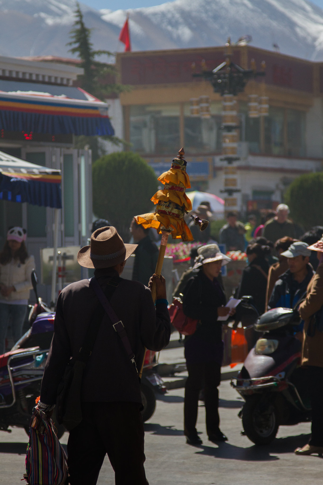 A man in a hat carries a saffron-draped prayer object through a busy Tibetan street below snow-capped mountains.