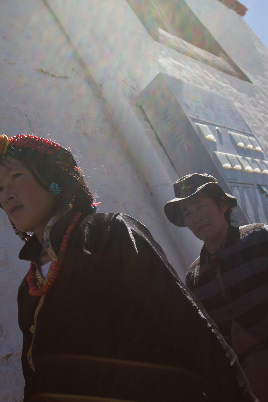 Two Tibetan pilgrims, a woman in coral beads and headdress and a man in a sun hat, beside a whitewashed wall.