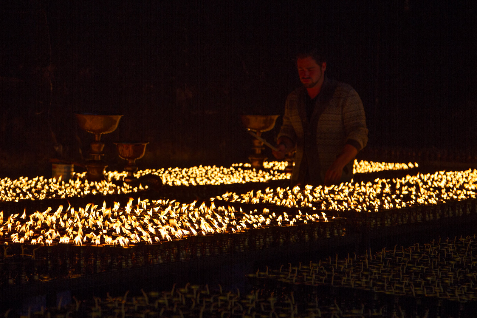A man tends rows of glowing butter lamps in a dark Tibetan monastery.