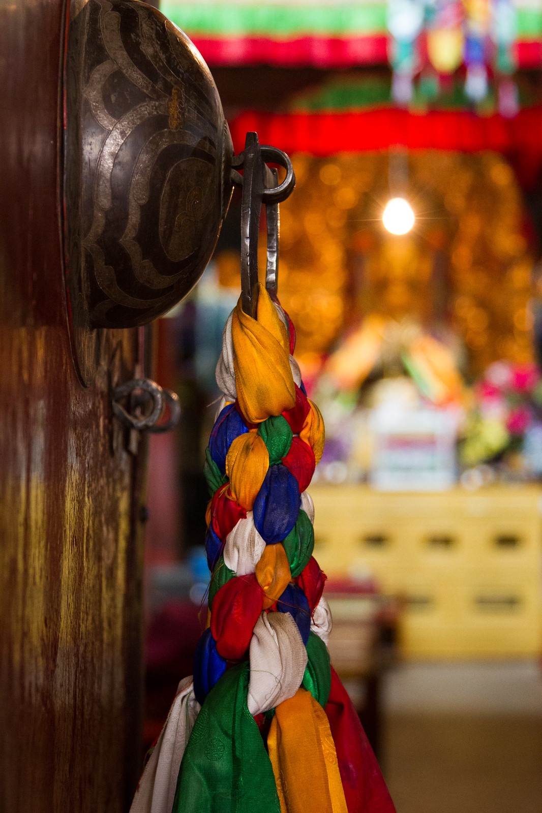 Braided colorful silk khata scarves hanging from an engraved bell inside a Tibetan Buddhist temple.