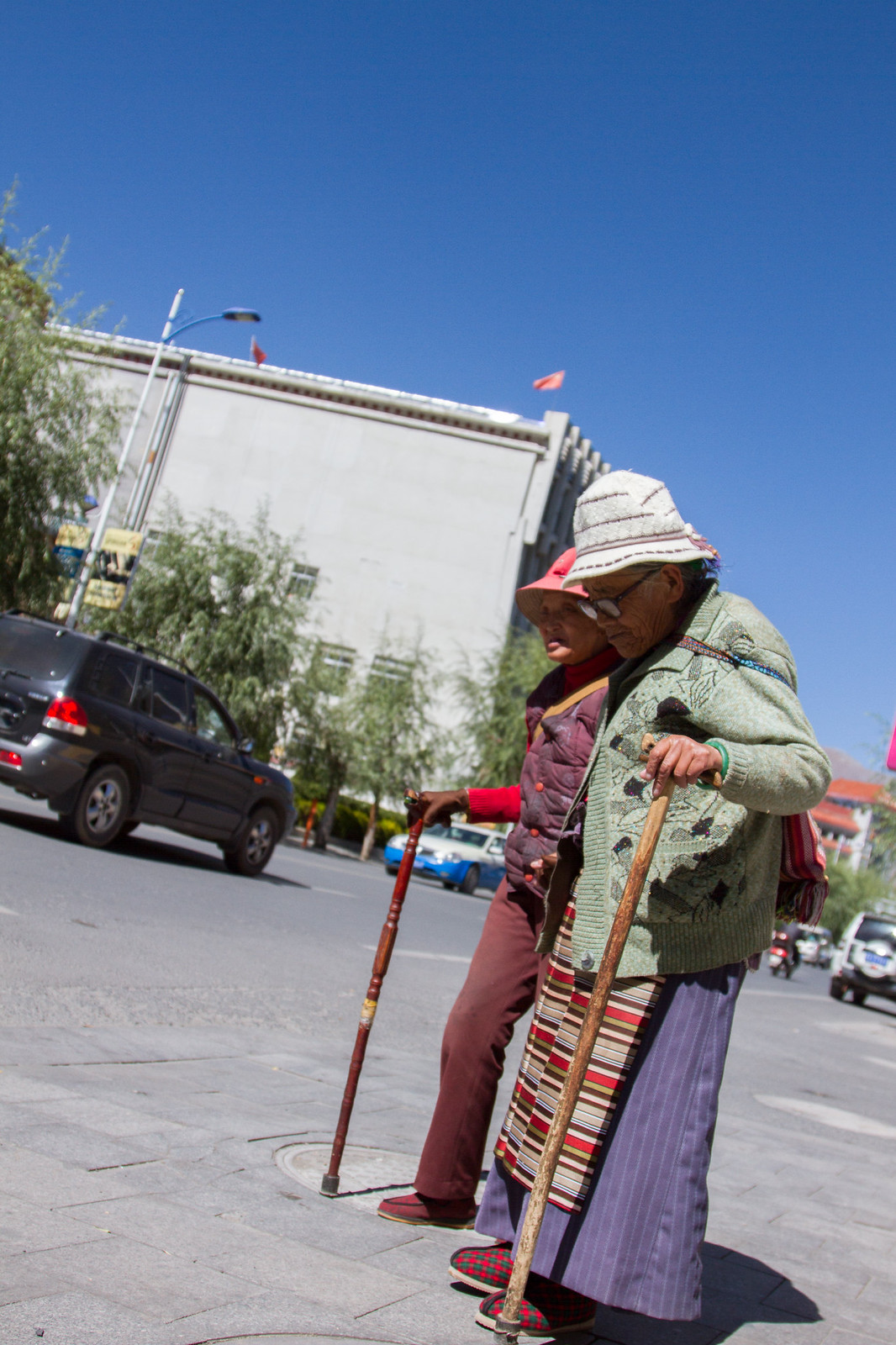 Two elderly Tibetan women in traditional dress lean on wooden canes crossing a sunny Lhasa street.