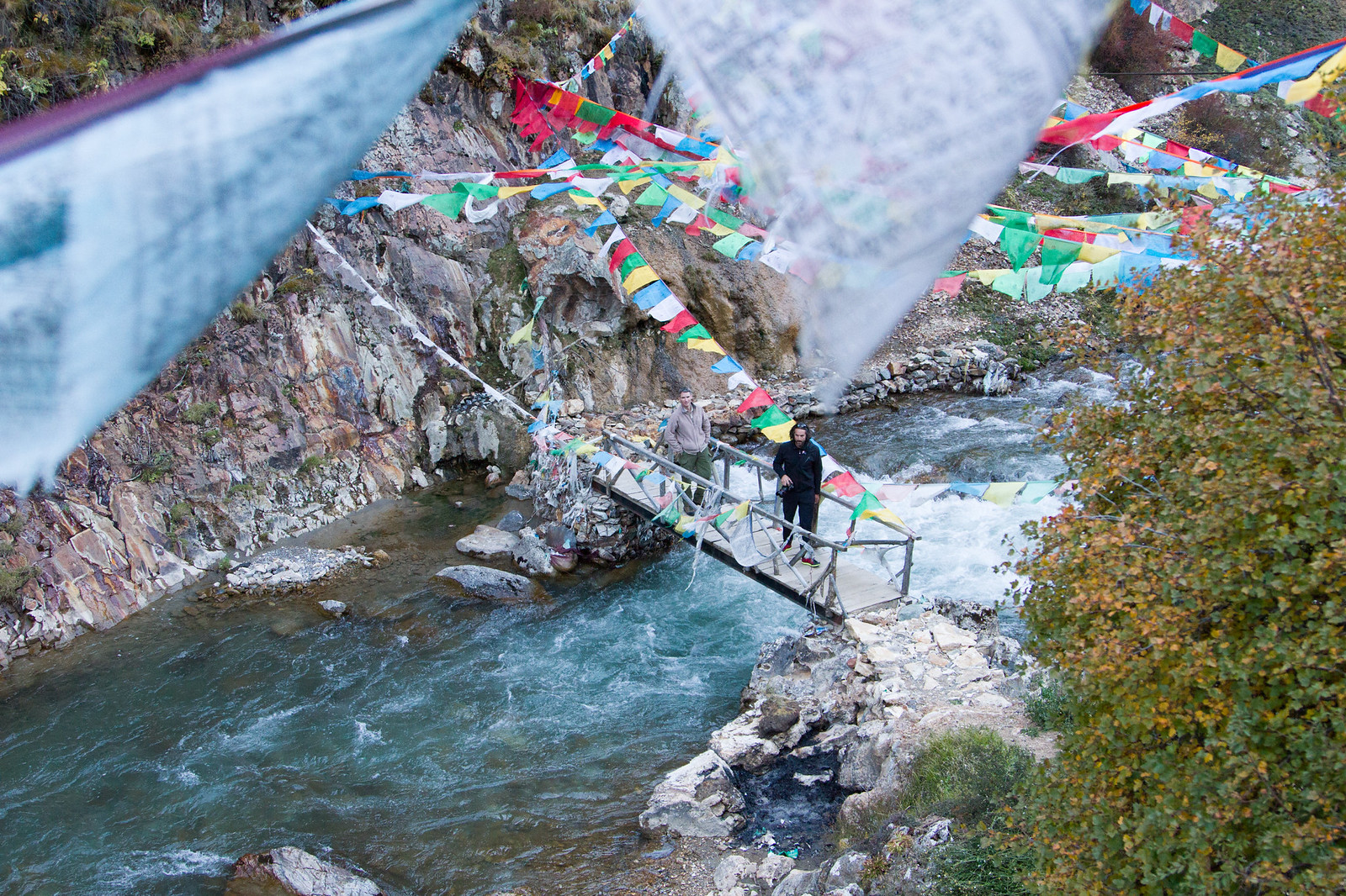 Two men cross a narrow wooden footbridge over a rushing mountain river strung with colorful Tibetan prayer flags.