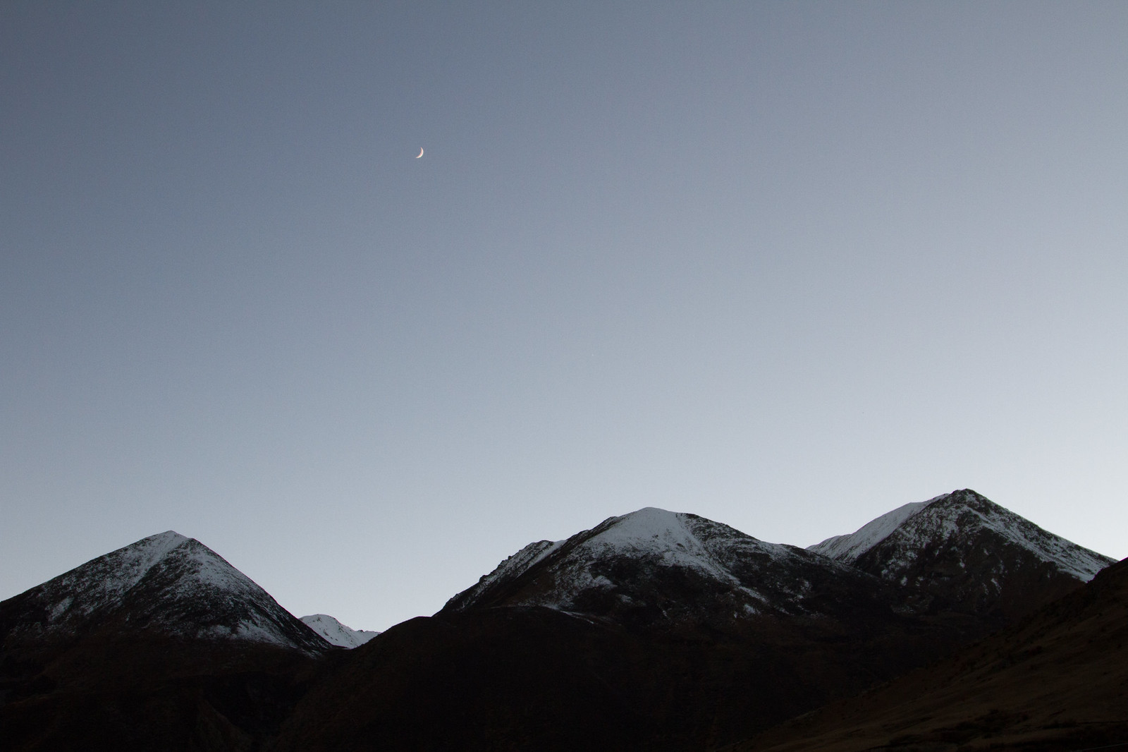 A crescent moon hangs over snow-dusted Tibetan mountain peaks in the fading blue light of dusk.