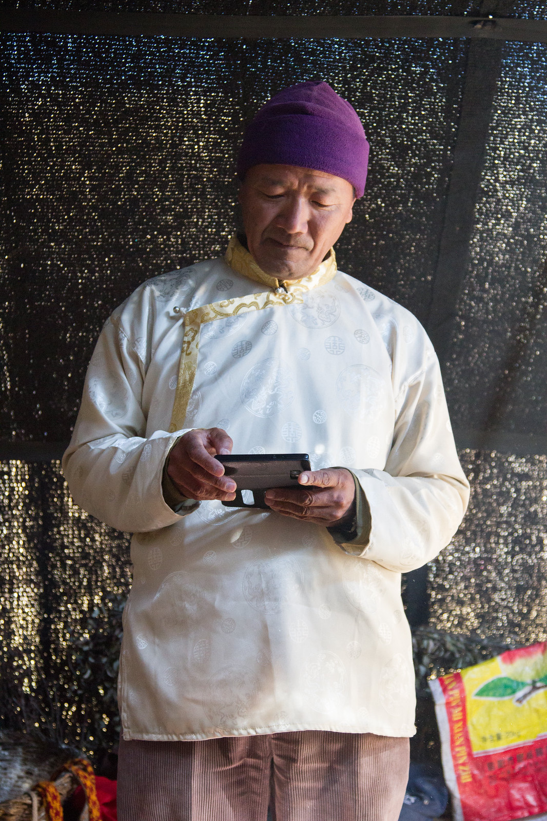 Man in a purple knit cap and cream silk Tibetan tunic reads a smartphone against a black mesh wall.