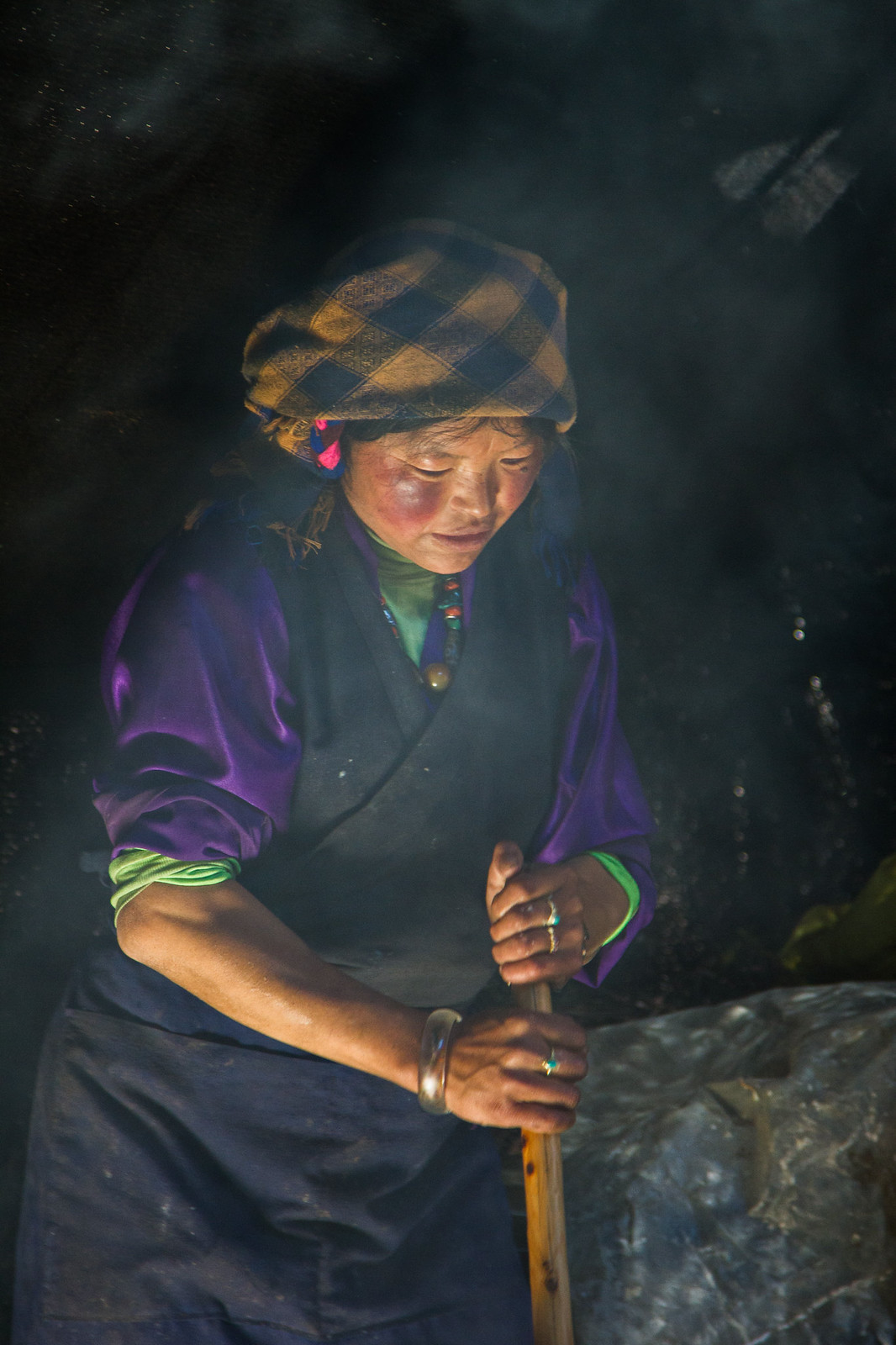 Tibetan woman in a purple robe and headscarf grips a wooden churn staff amid smoky, dim light.