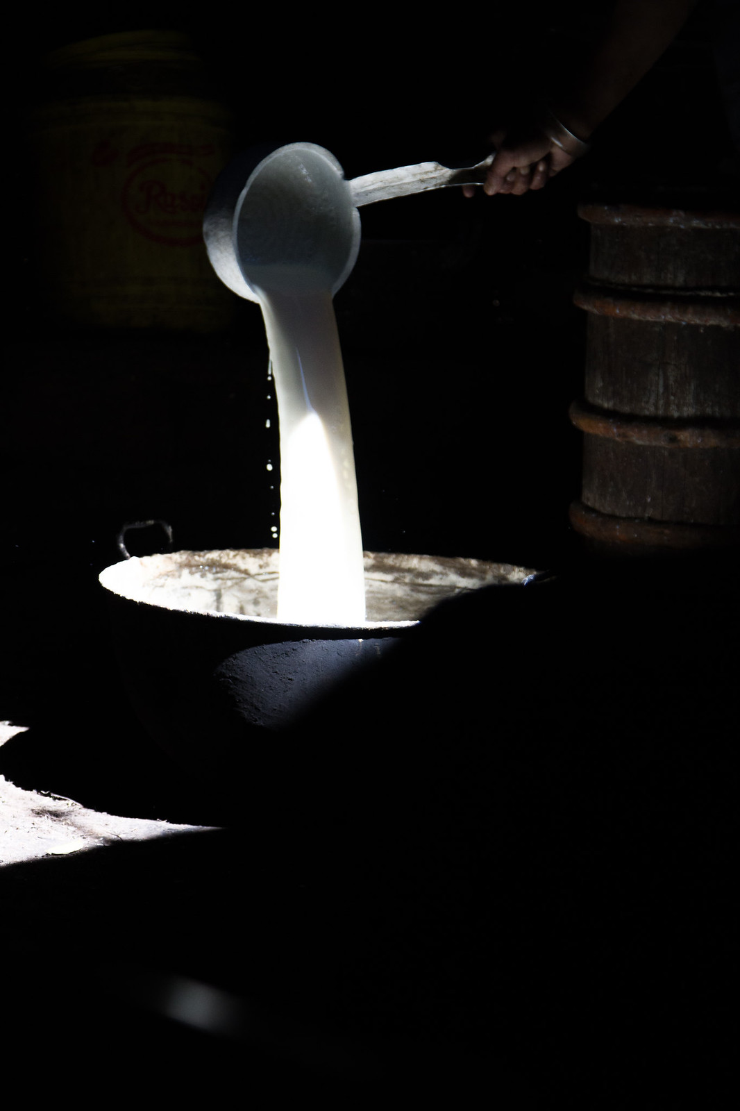 A hand pours milk from a ladle into a metal bowl in a dark room, beside a wooden churn.