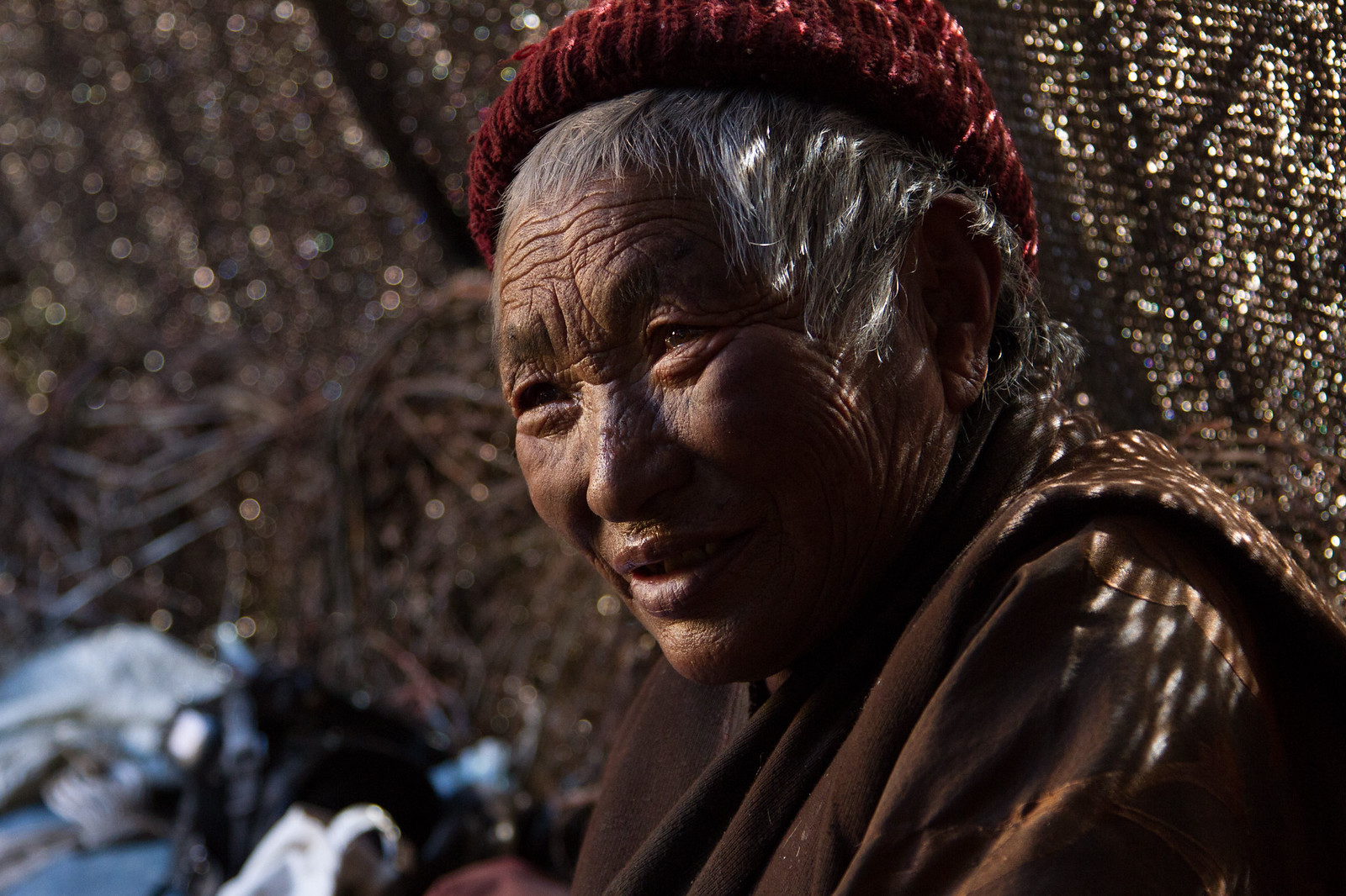 Elderly Tibetan woman in a red knit cap smiling in warm sunlight, her weathered face deeply lined.
