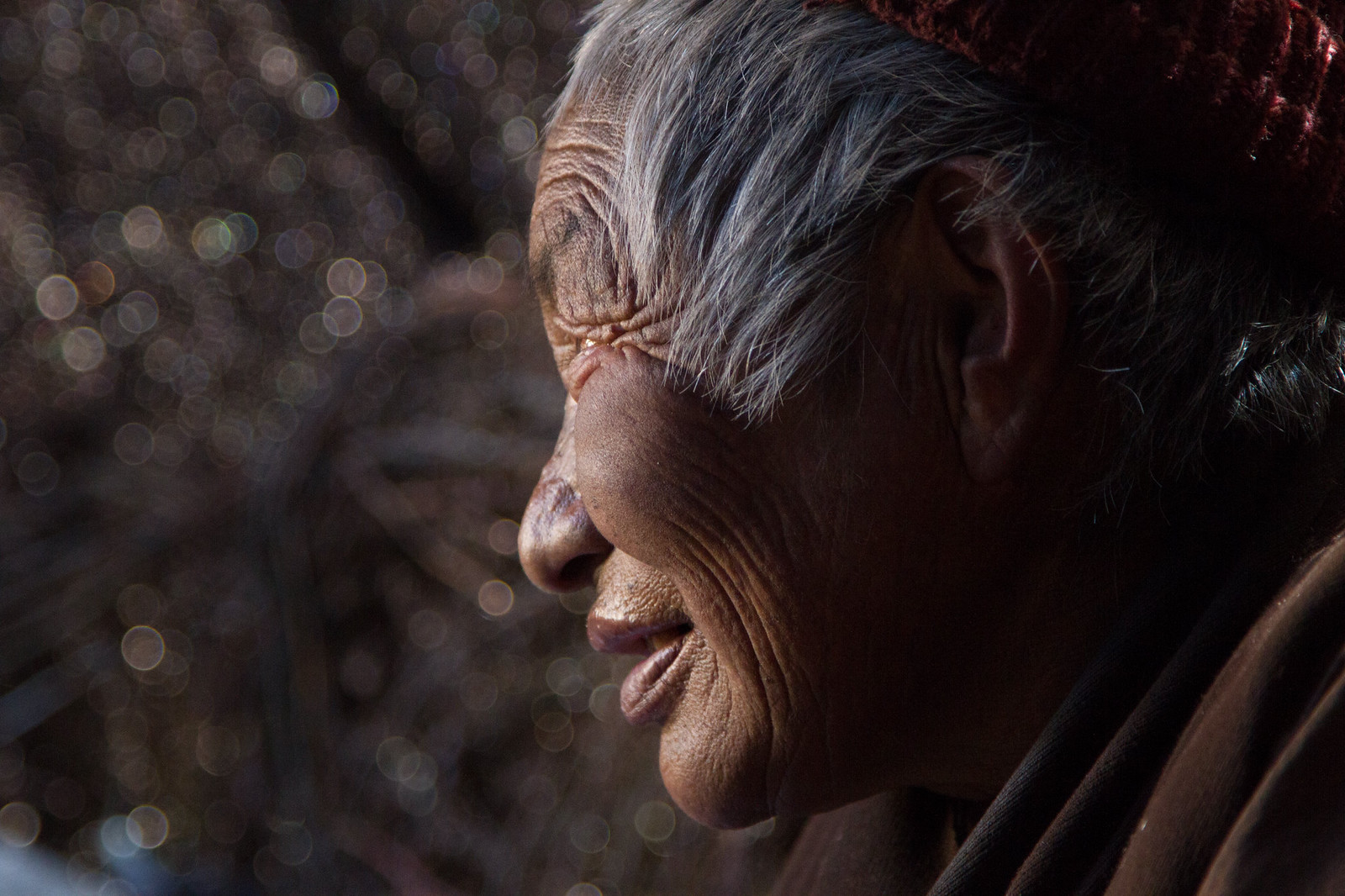 Weathered elderly Tibetan woman in profile, wearing a dark red knit cap, backlit by golden light.
