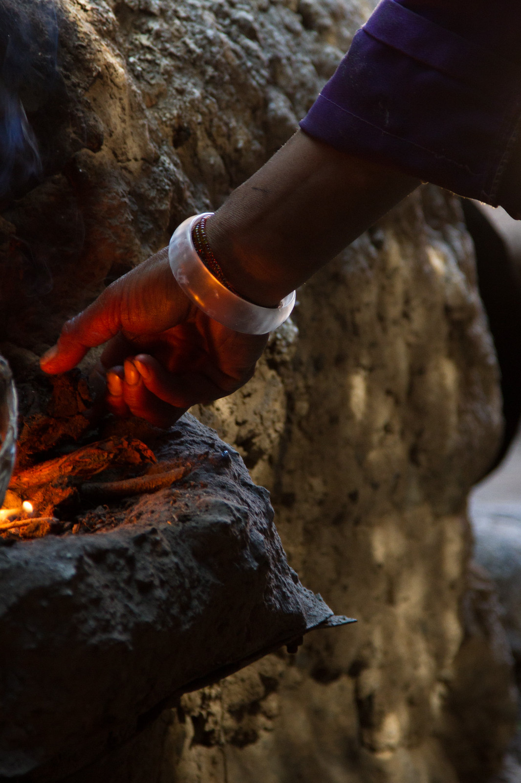 A hand with a beaded bracelet reaches down to tend glowing embers of a fire against dark rock.
