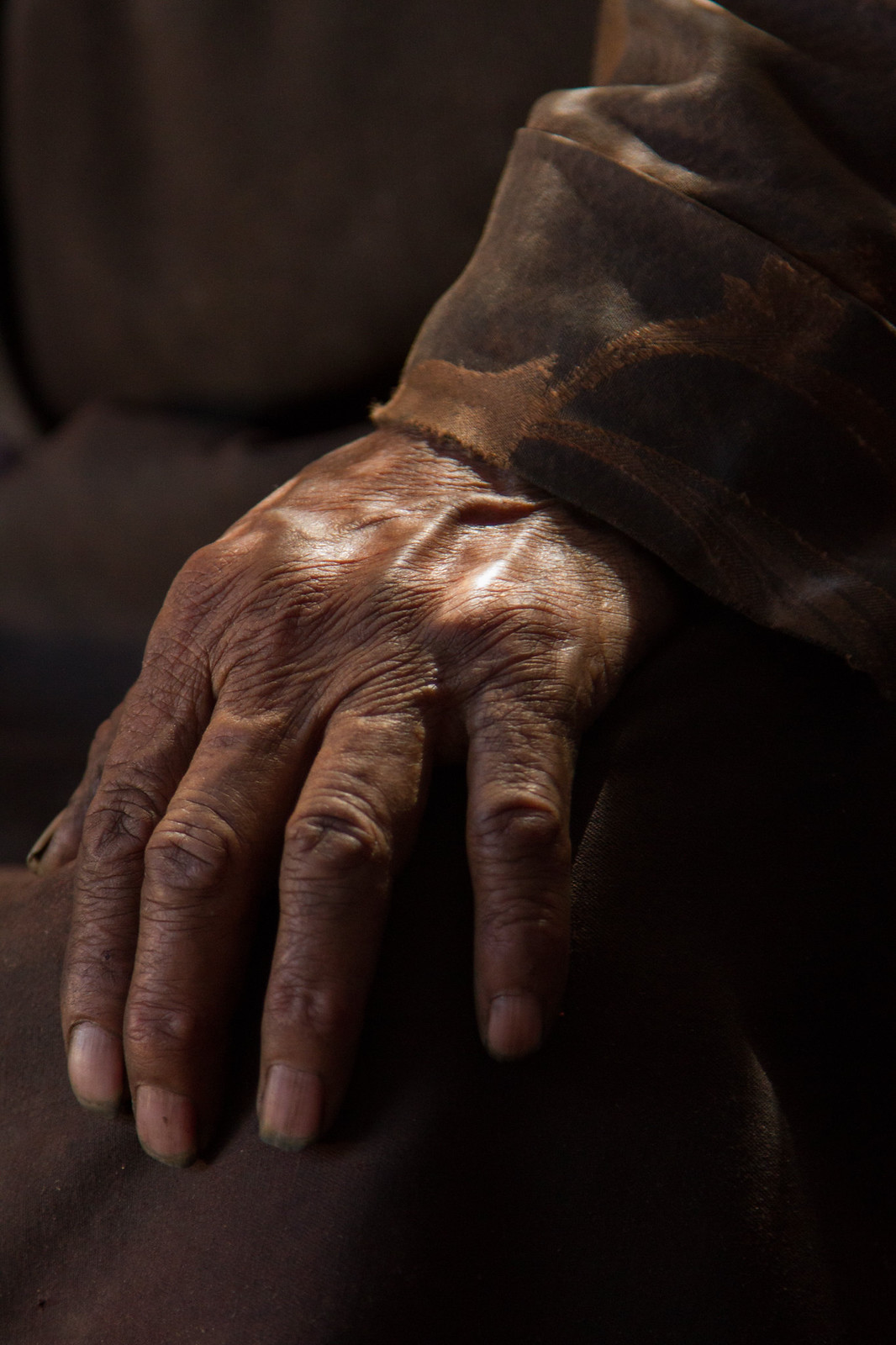 Weathered, deeply wrinkled hand of an elderly person resting on a knee, lit by warm side light against dark clothing.
