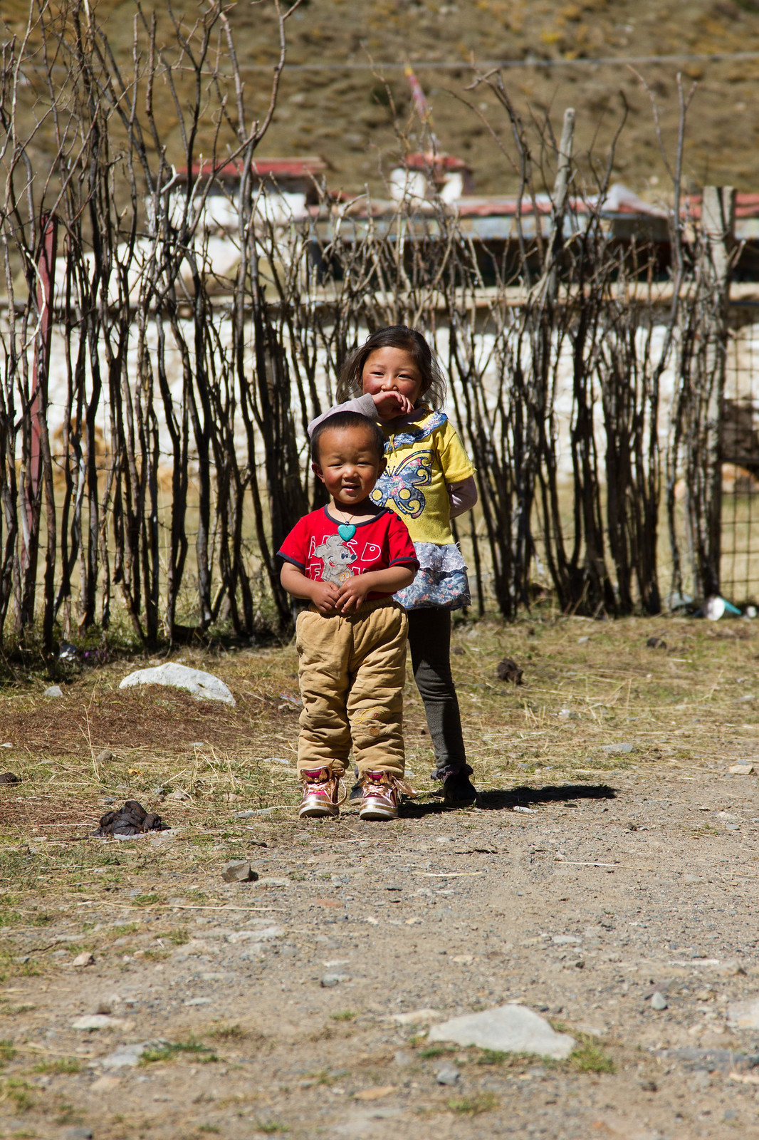 Two young Tibetan children stand on a dirt path by a wooden stick fence, one in a red shirt smiling at the camera.