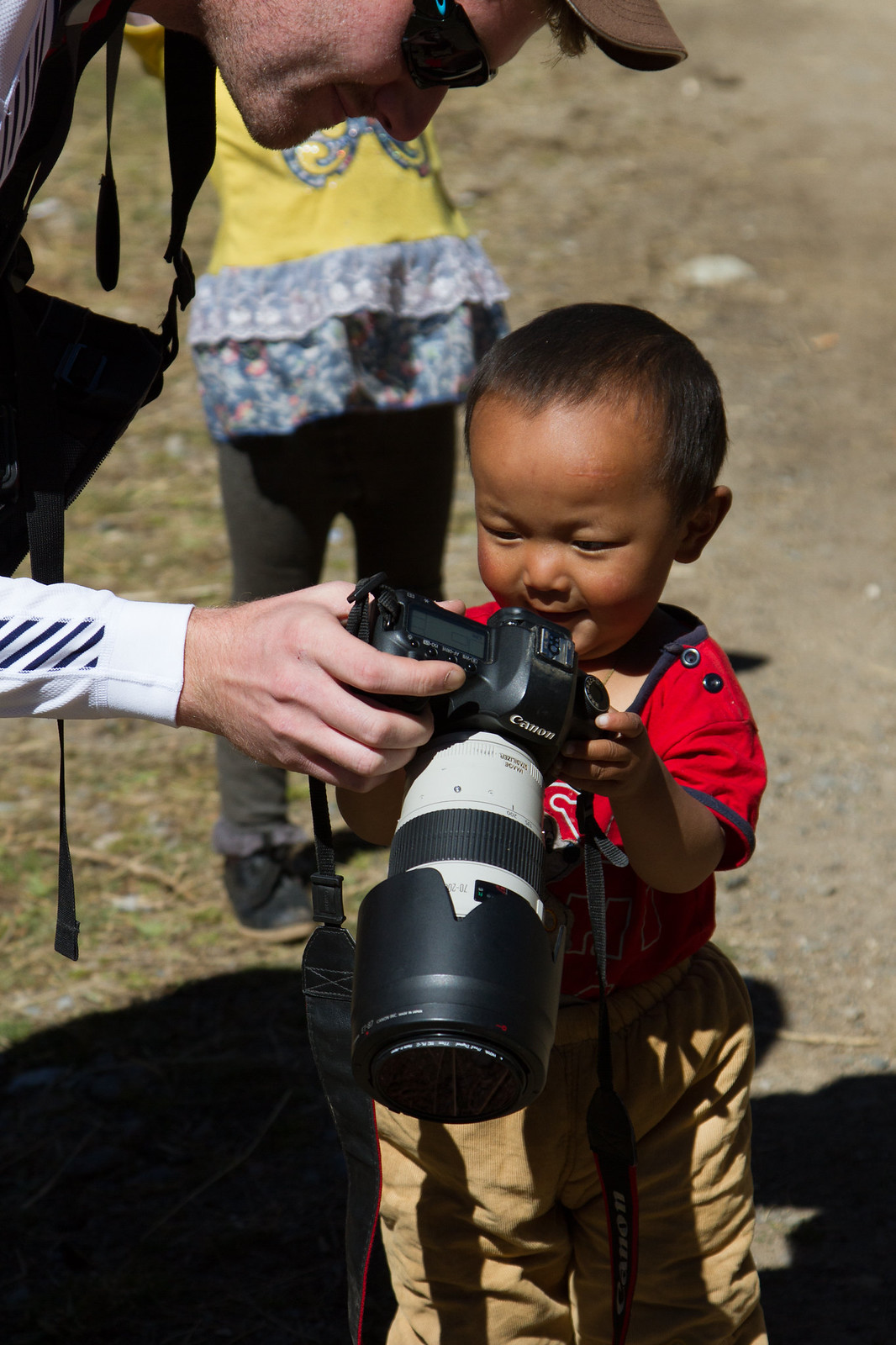 A young child in a red shirt peers into a large Canon DSLR camera held by a traveler outdoors.