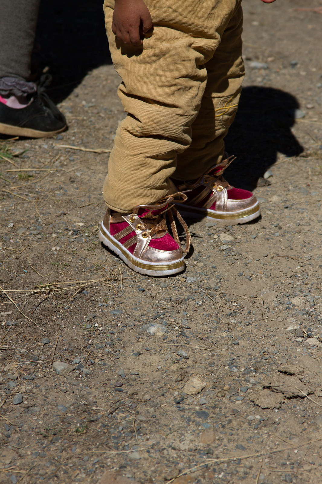 A child in tan corduroys and pink-and-gold sneakers stands on gravel beside an adult's foot.