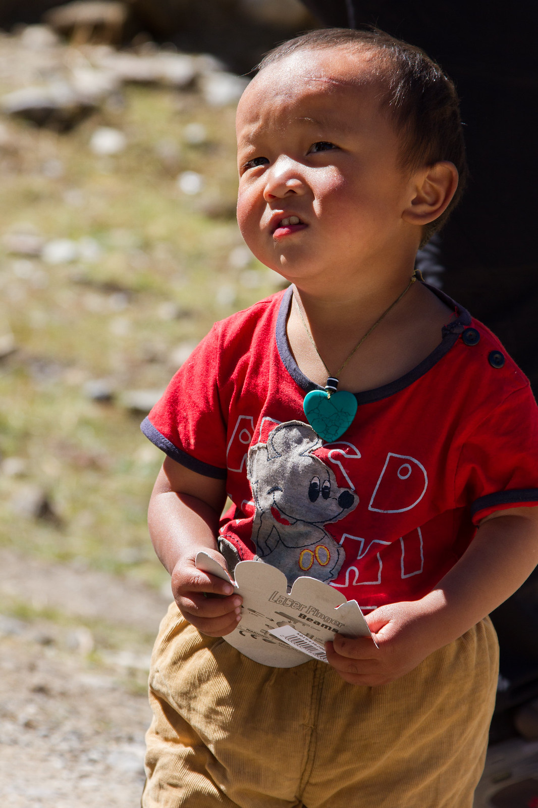 Young child in a red mouse-print T-shirt with a turquoise pendant, holding a "Laser Finger Beams" package outdoors.