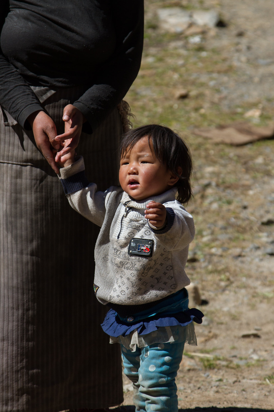 A young Tibetan child holds an adult's hand outdoors on a dusty, sunlit path.