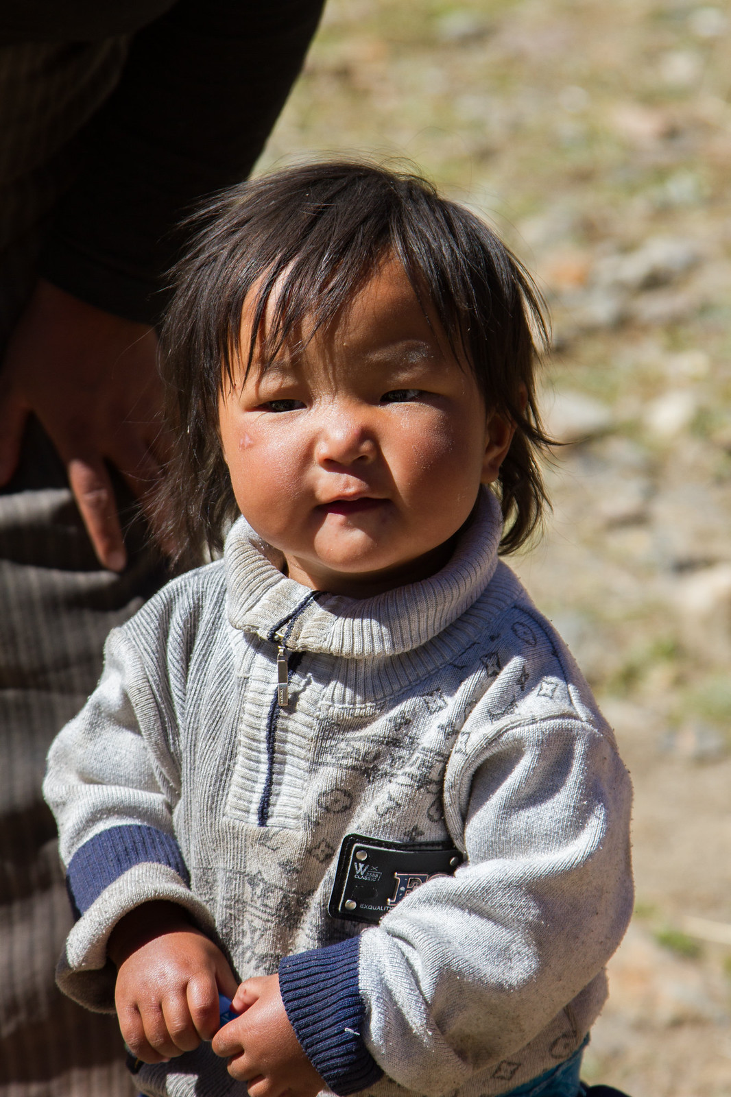 A young Tibetan child in a grey patterned sweater stands in sunlight beside an adult.