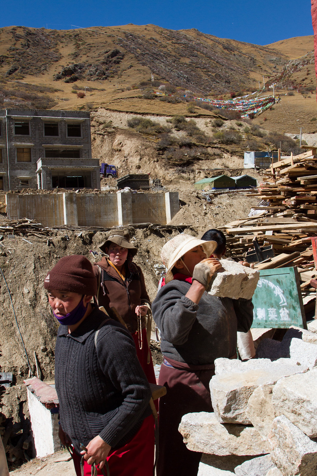 Tibetan workers, one shouldering a heavy stone block, at a hillside construction site with lumber piles and prayer flags.