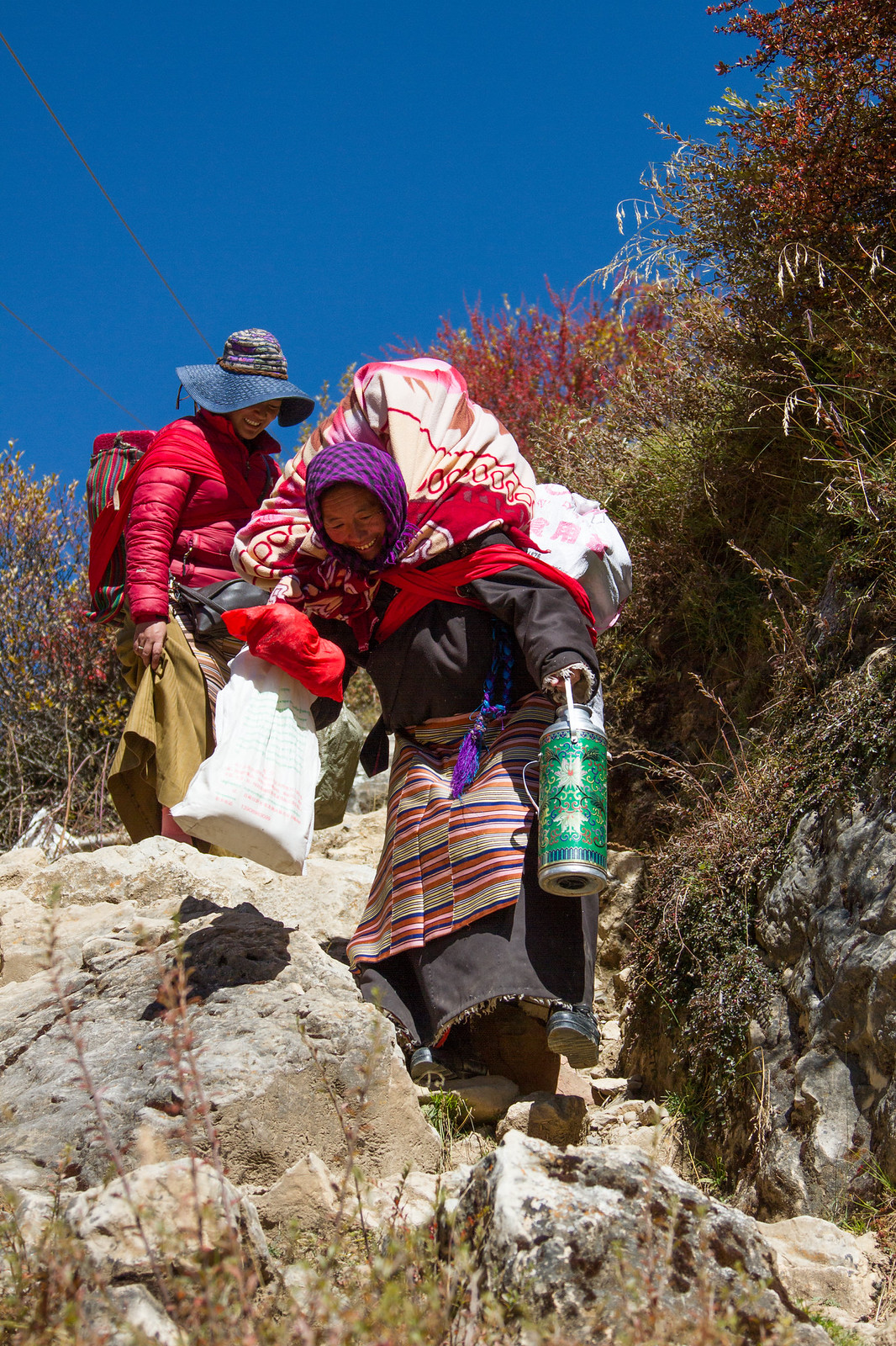 Two smiling Tibetan women in traditional dress climb a rocky trail carrying bundles under a bright blue sky.
