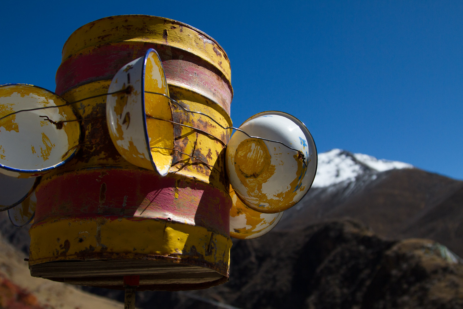 Weathered yellow and red oil drum topped with painted enamel bowls against a snow-capped Tibetan peak