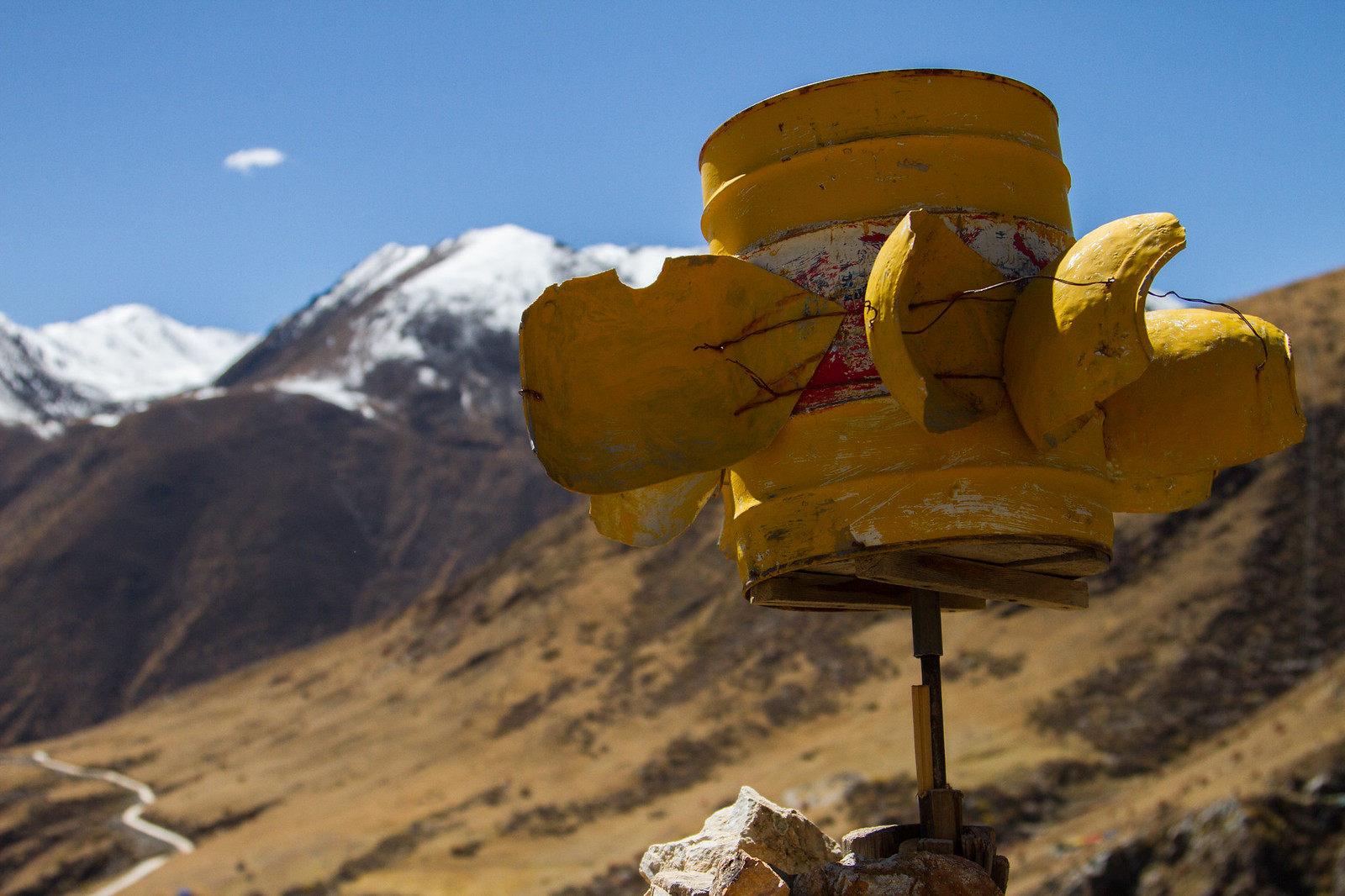 A yellow makeshift wind turbine made from a cut oil drum against snow-capped Tibetan mountains and a winding valley road.