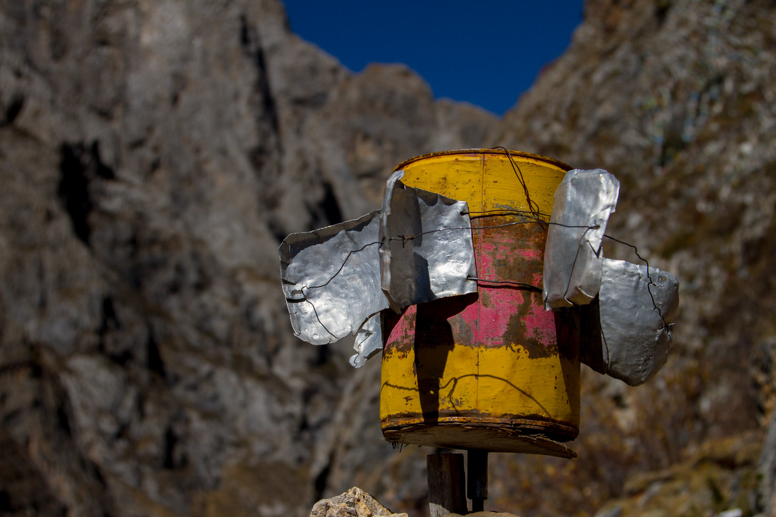 A weathered yellow bucket rigged with scrap-metal blades as a makeshift wind spinner against rocky Tibetan mountains.