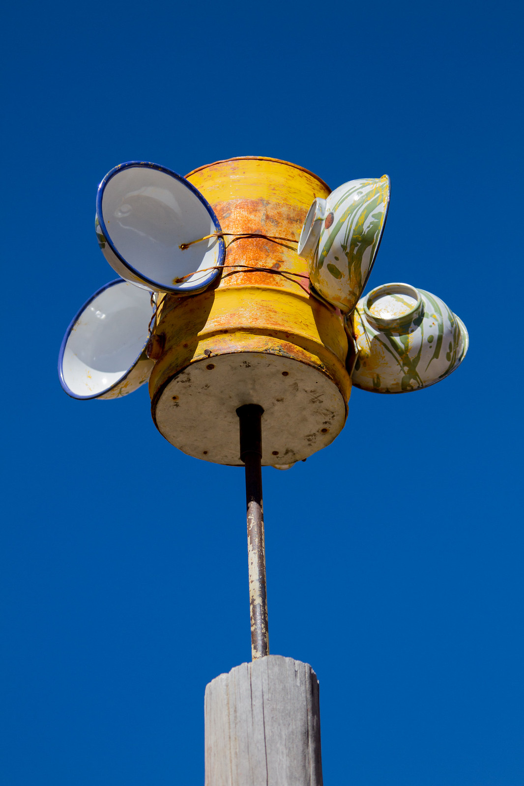 A rusty yellow tin with enamel bowls wired to it atop a wooden post against deep blue sky.