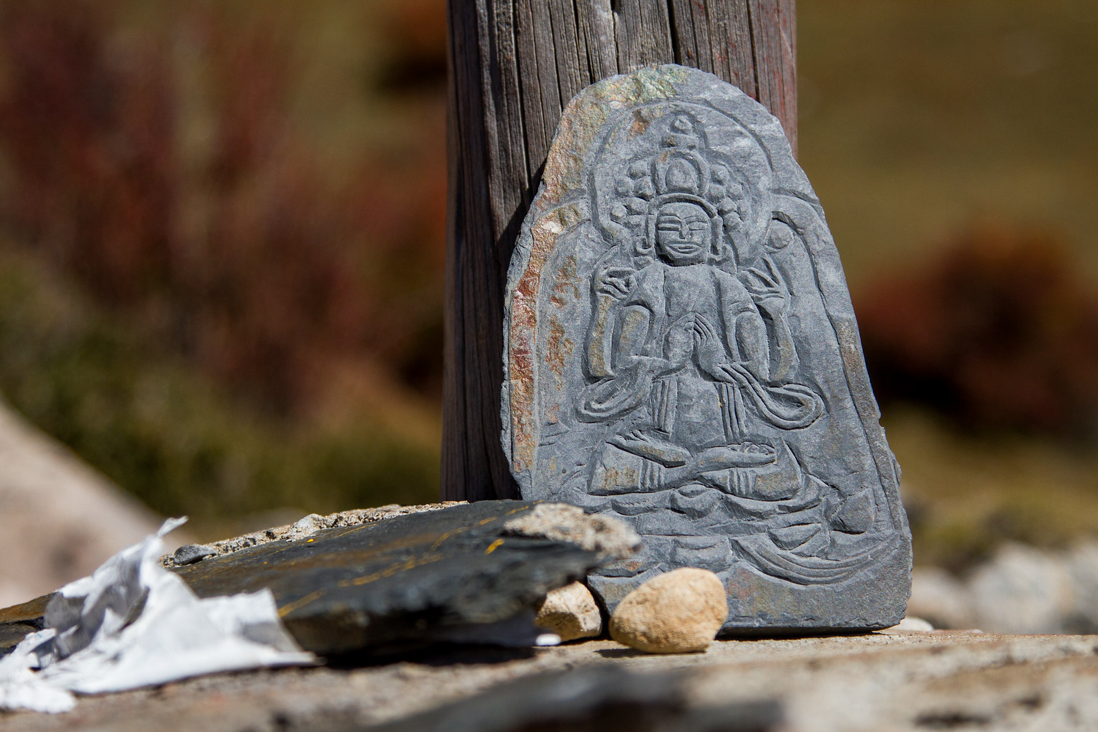 A carved stone mani slab depicting a seated Buddha leaning against a weathered wooden post outdoors.
